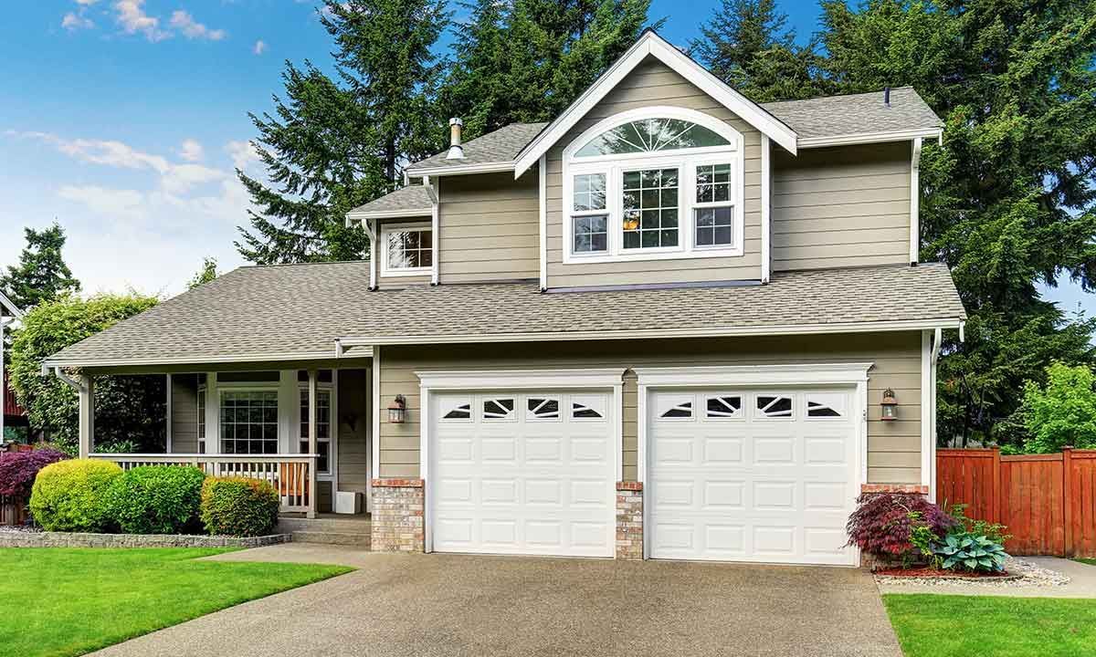 Two-story house with two-car garage, beige siding, gray roof, green lawn, trees, and landscaping.
