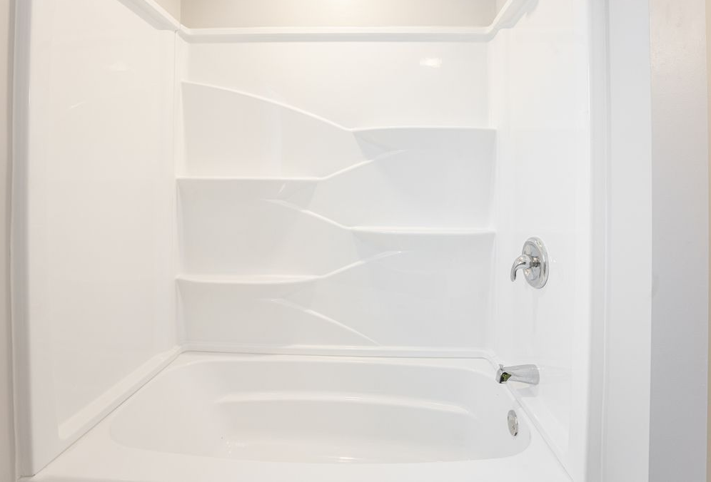 White, built-in bathtub and shower unit. Three tiered shelves on back wall, chrome fixtures.