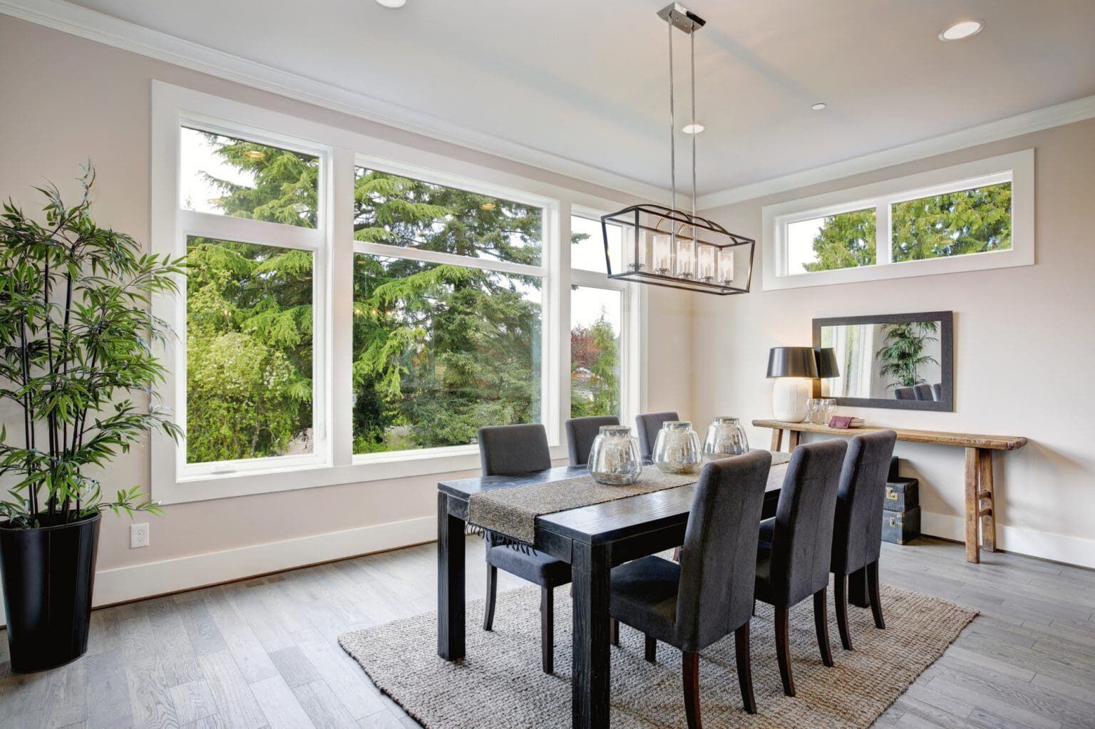 Dining room with large windows, dark table and chairs, rug, and greenery; natural light.