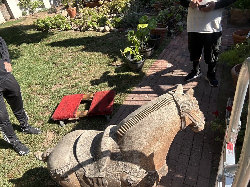 A stone horse statue being moved outdoors by two people. Red dolly and greenery in the background.