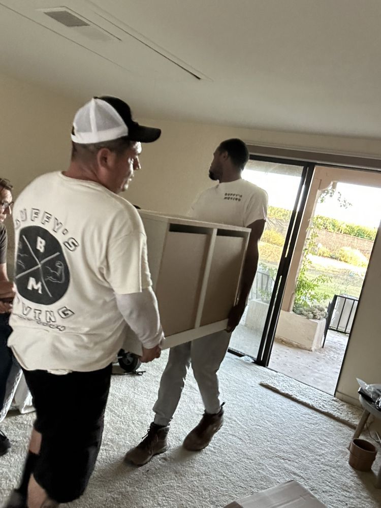 Two people carrying a light-colored cabinet through a doorway, likely moving furniture.
