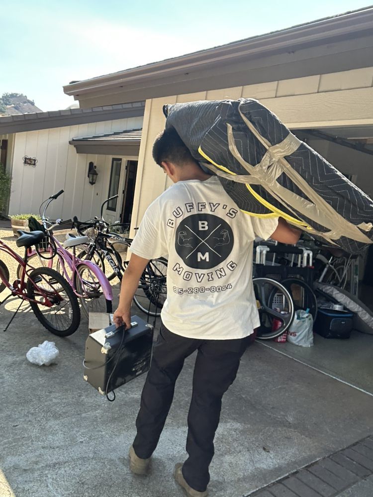 Person carrying a large bag over shoulder and a box, near bikes and a garage.