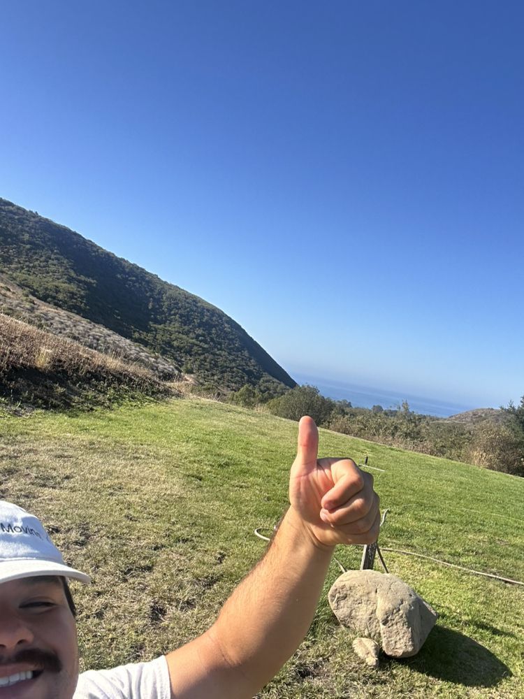 Man giving thumbs up on a grassy hill, with mountain and ocean views under a blue sky.
