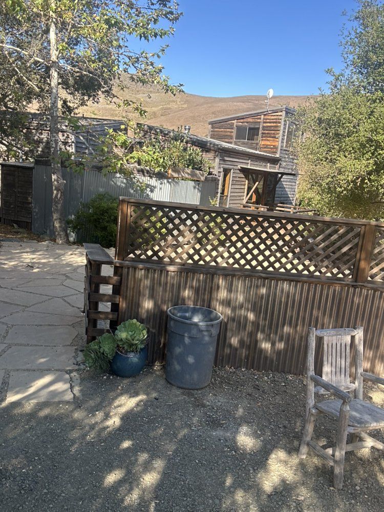 Wooden fence and shed in a yard, blue trash can, potted plants, clear sky.