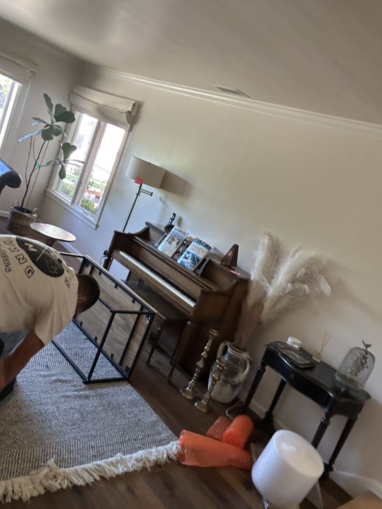 Person bending over a black frame on a rug, piano, and side table in a room.