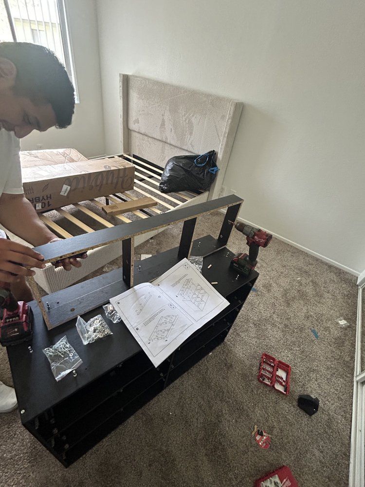 Person assembling a black shelving unit with instructions and a drill in a bedroom.