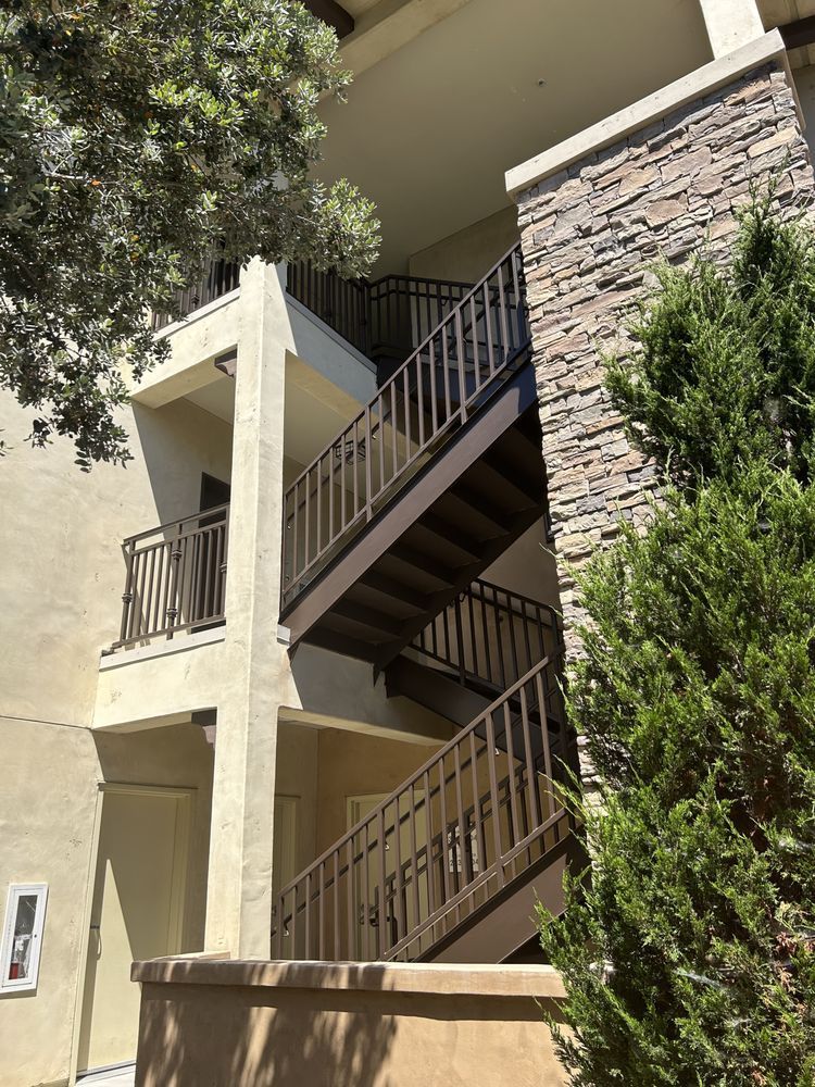 Exterior stairs of a building with brown railings and stone accents, flanked by greenery and trees.