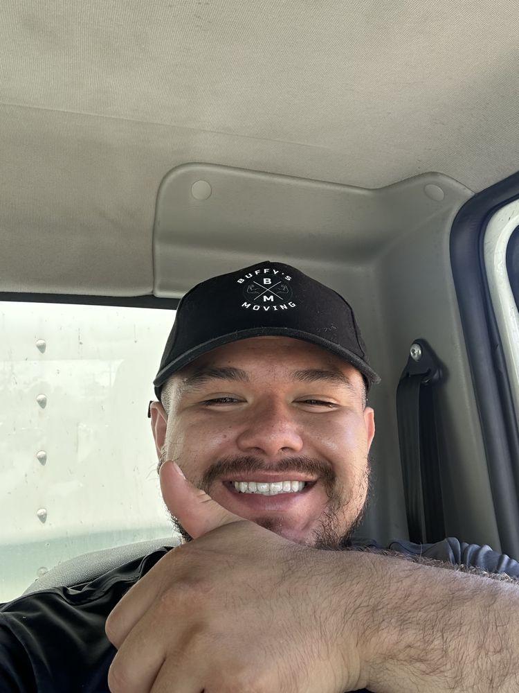 Man in black cap smiles, gives thumbs up, inside vehicle.