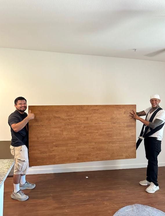 Two people holding a large, brown wooden board inside a room. One gives a thumbs-up.