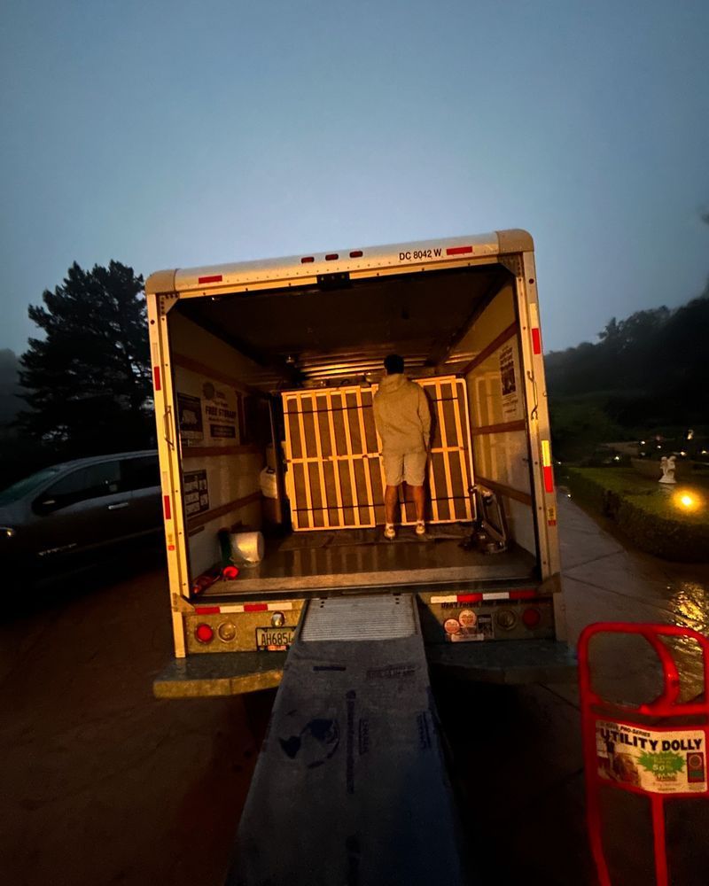 A person loads a wooden crate into a moving truck, outdoors, on a cloudy day.