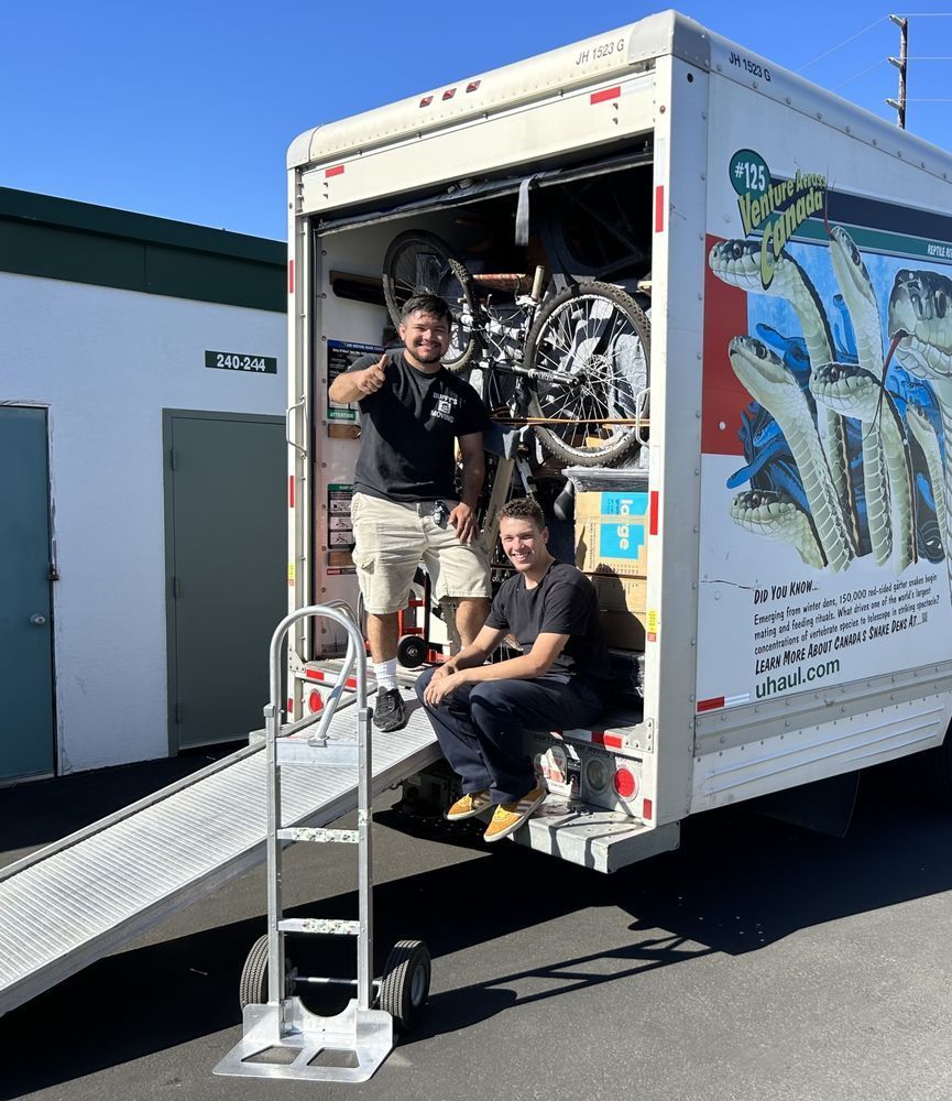 Two men with a moving truck, bikes inside, and a hand truck on ramp at a storage unit. One gives a thumbs-up.