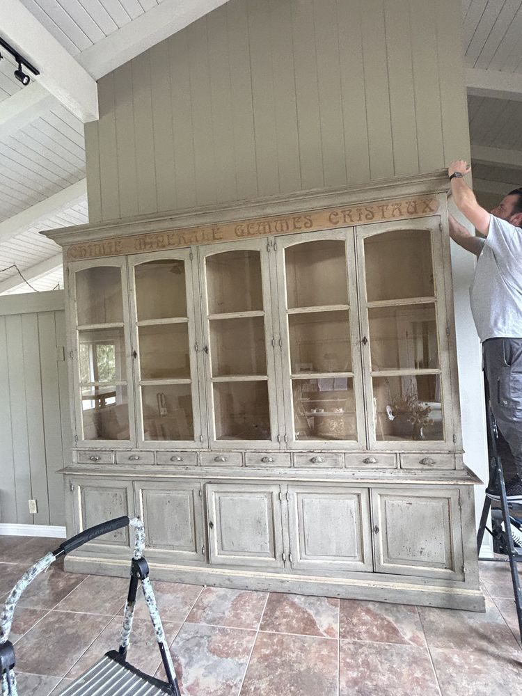 A person installing a large, antique, gray cabinet with glass doors on a wall.