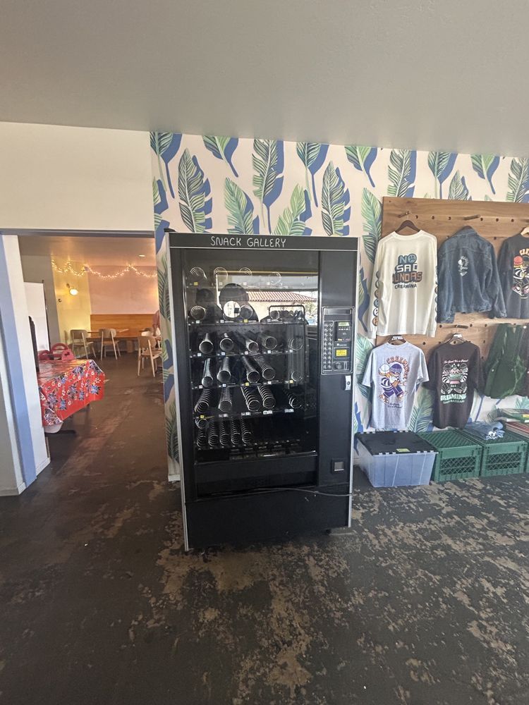 Black vending machine in a shop with t-shirts and a dining area visible.