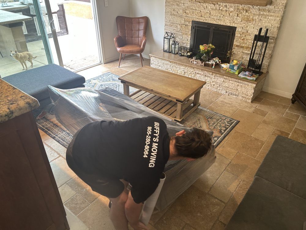 Person moving a large, wrapped furniture piece in a living room. Fireplace, table, and rug are visible.