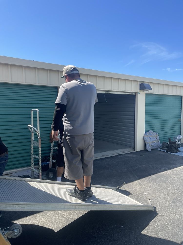 Man on ramp, looking into an open storage unit. Hand truck and another person present. Bright, sunny day.