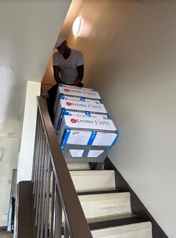 Person carrying three large boxes up a stairwell. Boxes are white, blue, and red. Person wears a white hat and grey shirt.