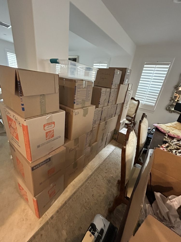 Boxes stacked in a room, partially blocking a doorway, next to dining chairs and a window.