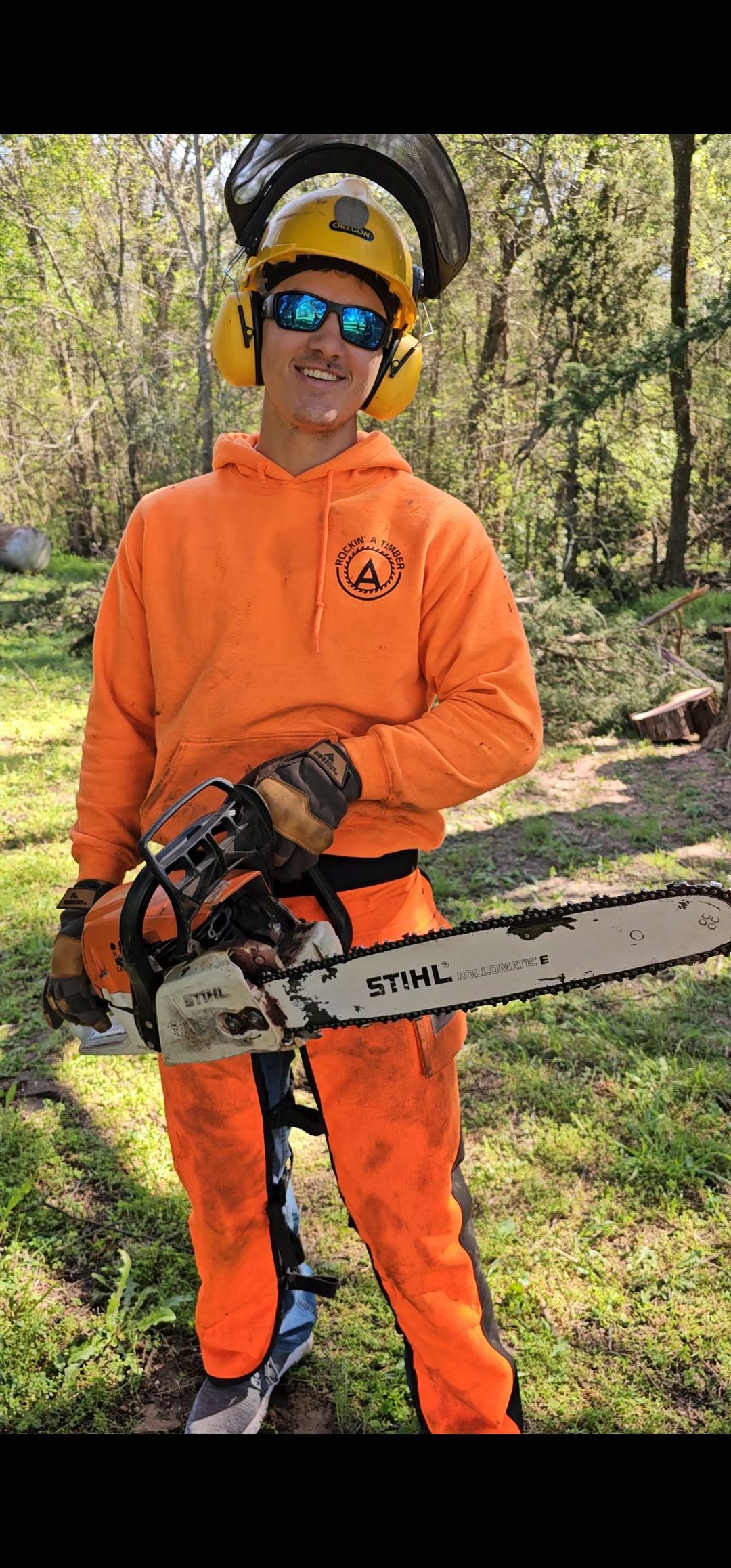 Person in orange work attire holding a chainsaw, wearing ear and eye protection, standing outdoors.