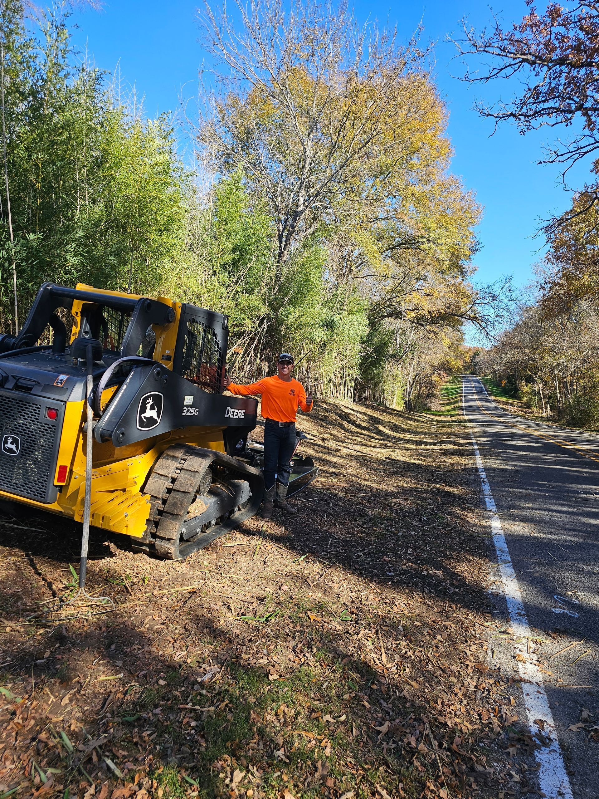 Person operating a yellow skid steer clears brush alongside a road with fall foliage.