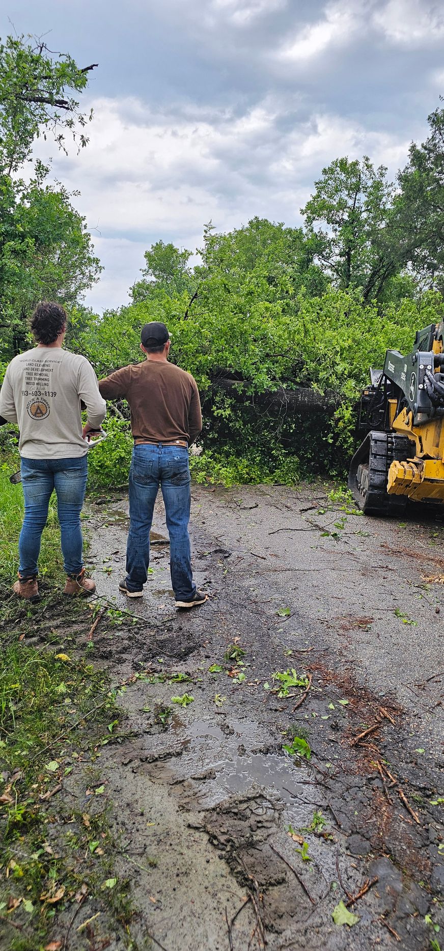 Two men in jeans observe a tree blocking a muddy road, a yellow machine on the right.