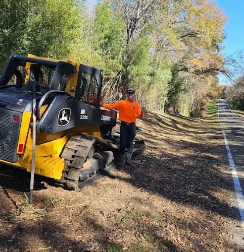 Man in orange shirt stands next to a John Deere skid steer on roadside. Trees and asphalt road in background.