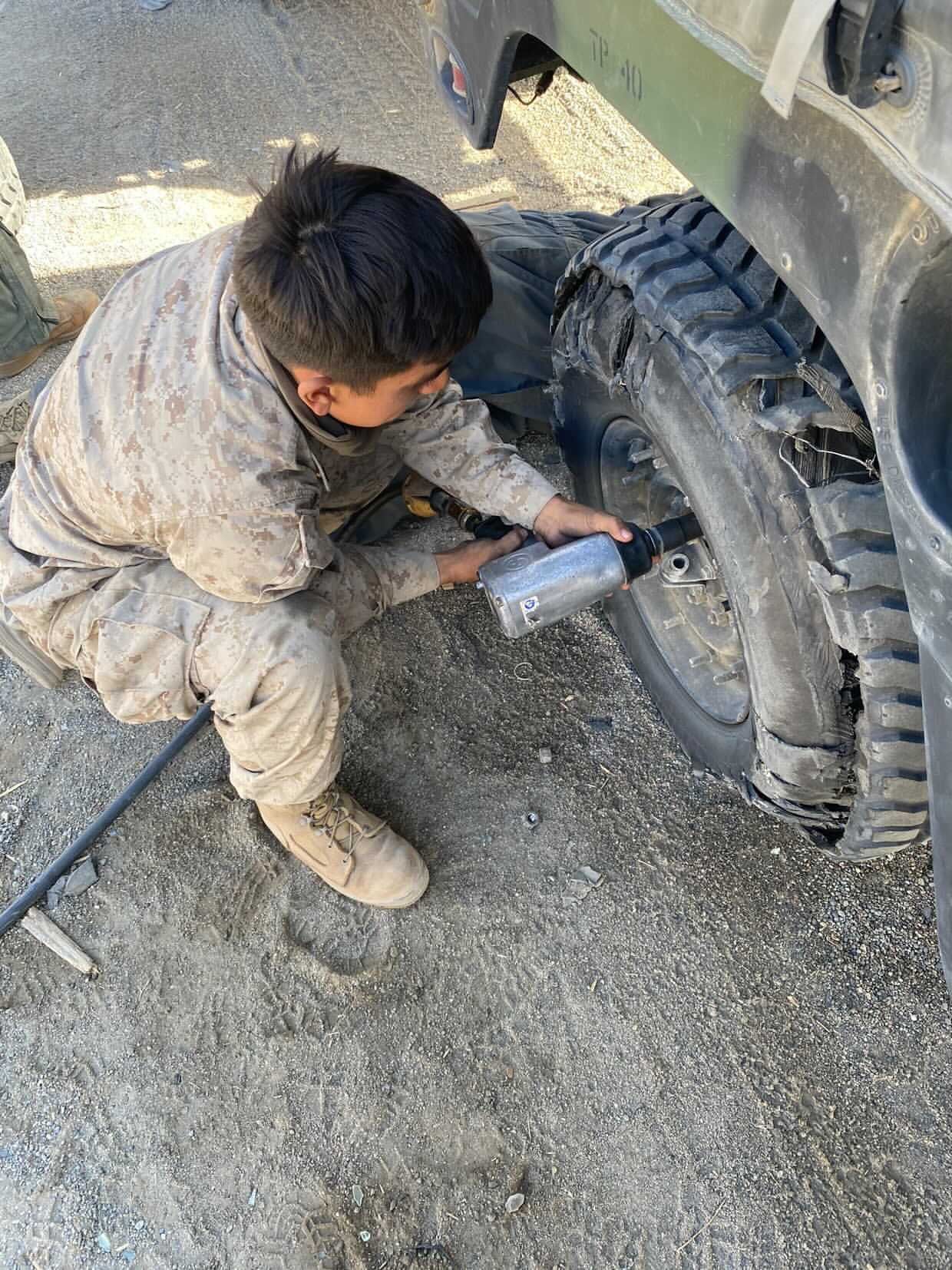 A person in camouflage uniform kneeling on dirt, using a power tool to remove a lug nut from a vehicle tire.