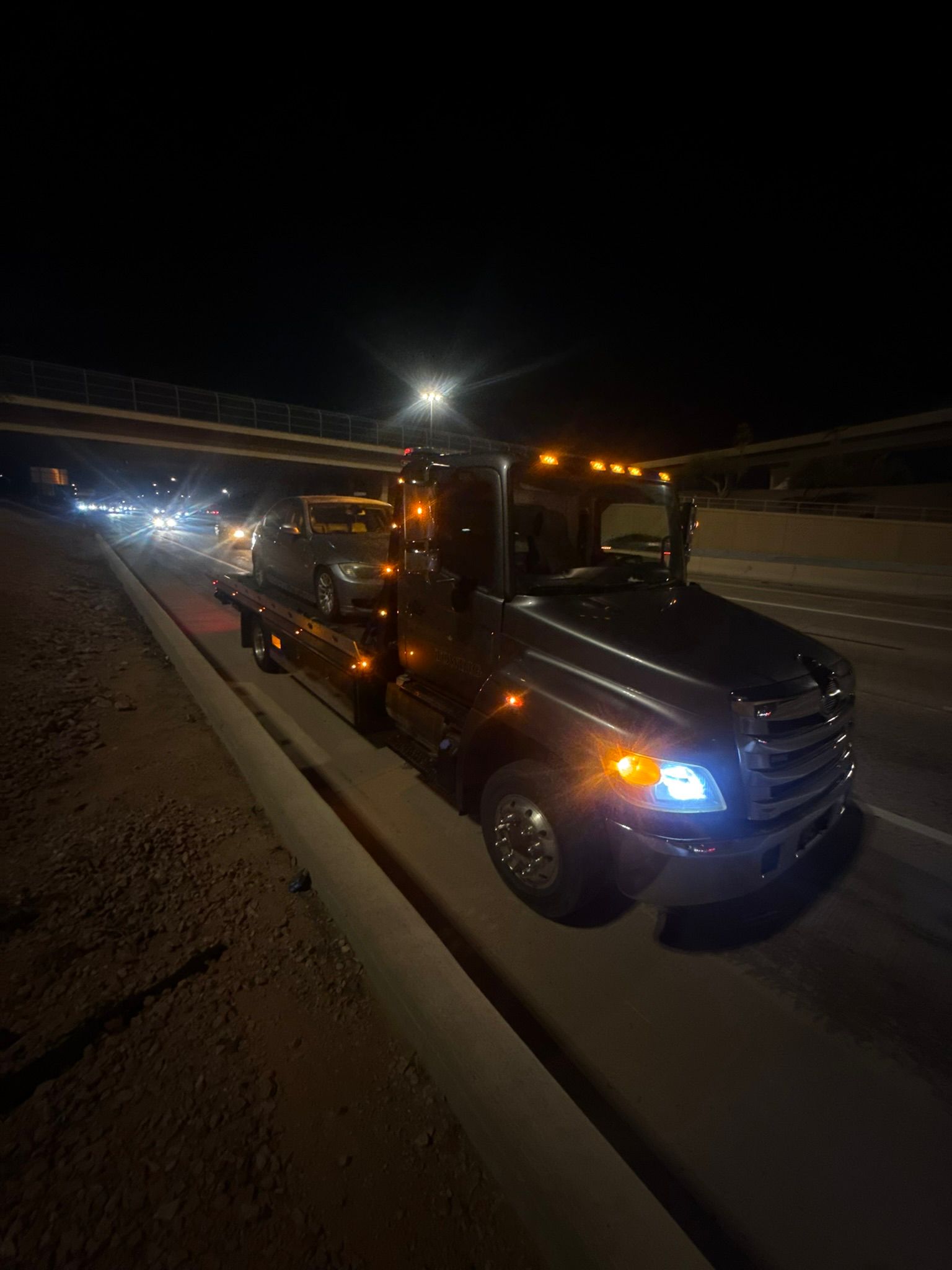 A tow truck with its headlights and orange marker lights on, carrying a car on its flatbed at night on a highway.