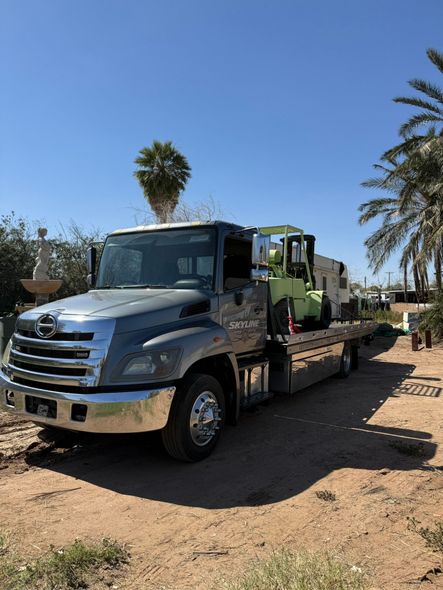 A gray flatbed tow truck carrying a light green forklift, parked on a dirt lot with palm trees in the background.