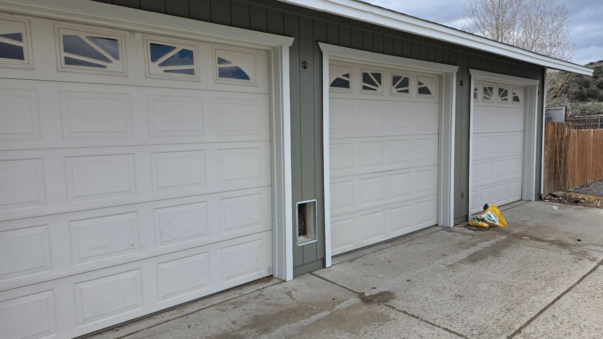 Three white garage doors on a gray driveway beside a house, with a small item near the right door.