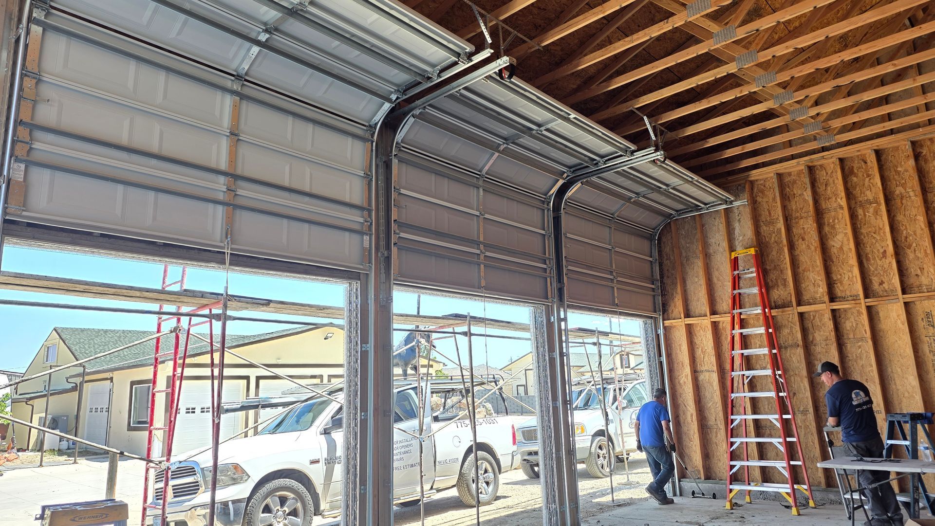 Interior of a garage under construction with open doors, exposed framing, ladders, and workers near vehicles outside