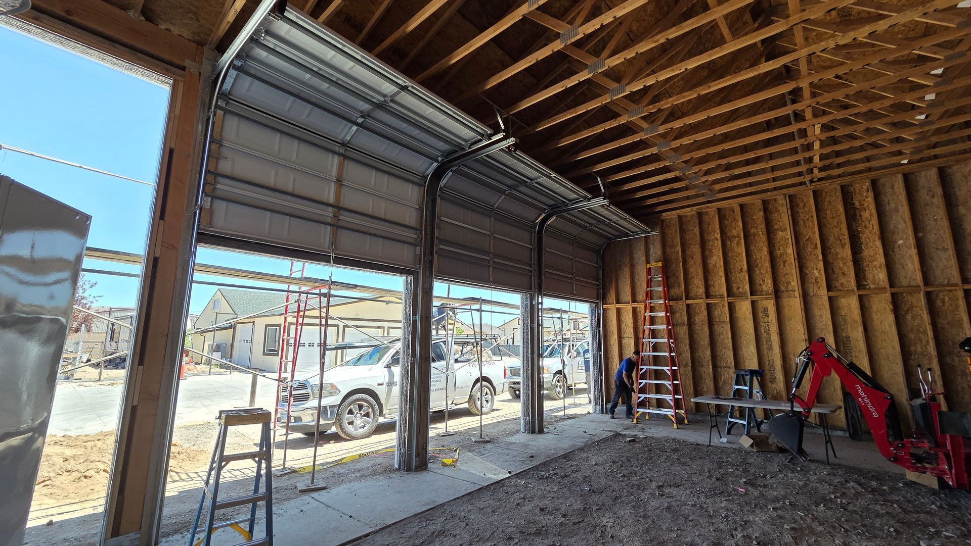 Construction site interior with open garage door, exposed framing, ladders, and a red excavator near the wall