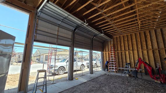 Construction site interior with open garage door, exposed framing, ladders, and a red excavator near the wall