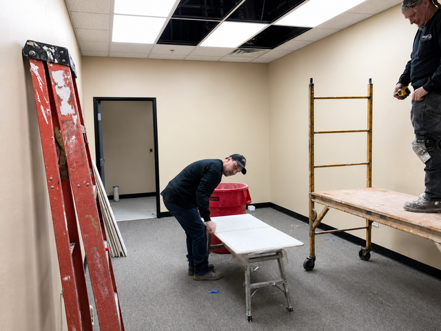 Two workers moving materials in a room with a scaffold and ladder under ceiling repair.