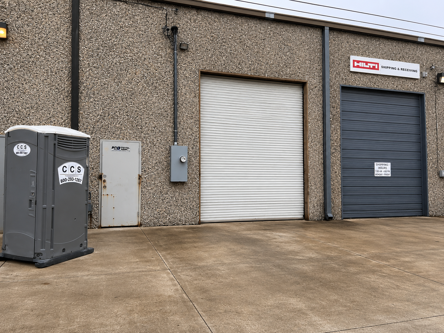 Industrial building exterior with two roll-up doors, portable toilet, and concrete lot