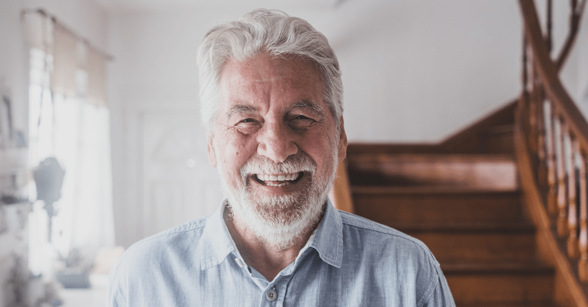 An elderly man with a beard is smiling in front of a wooden staircase.