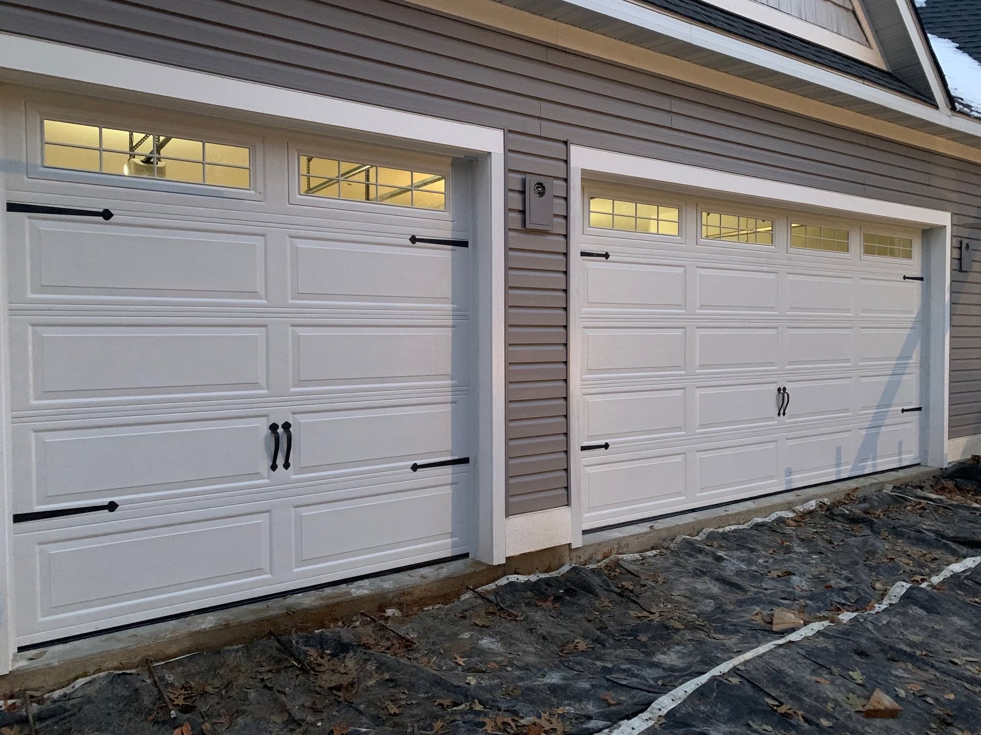 Two white garage doors on a house exterior with small windows and unfinished ground in front.