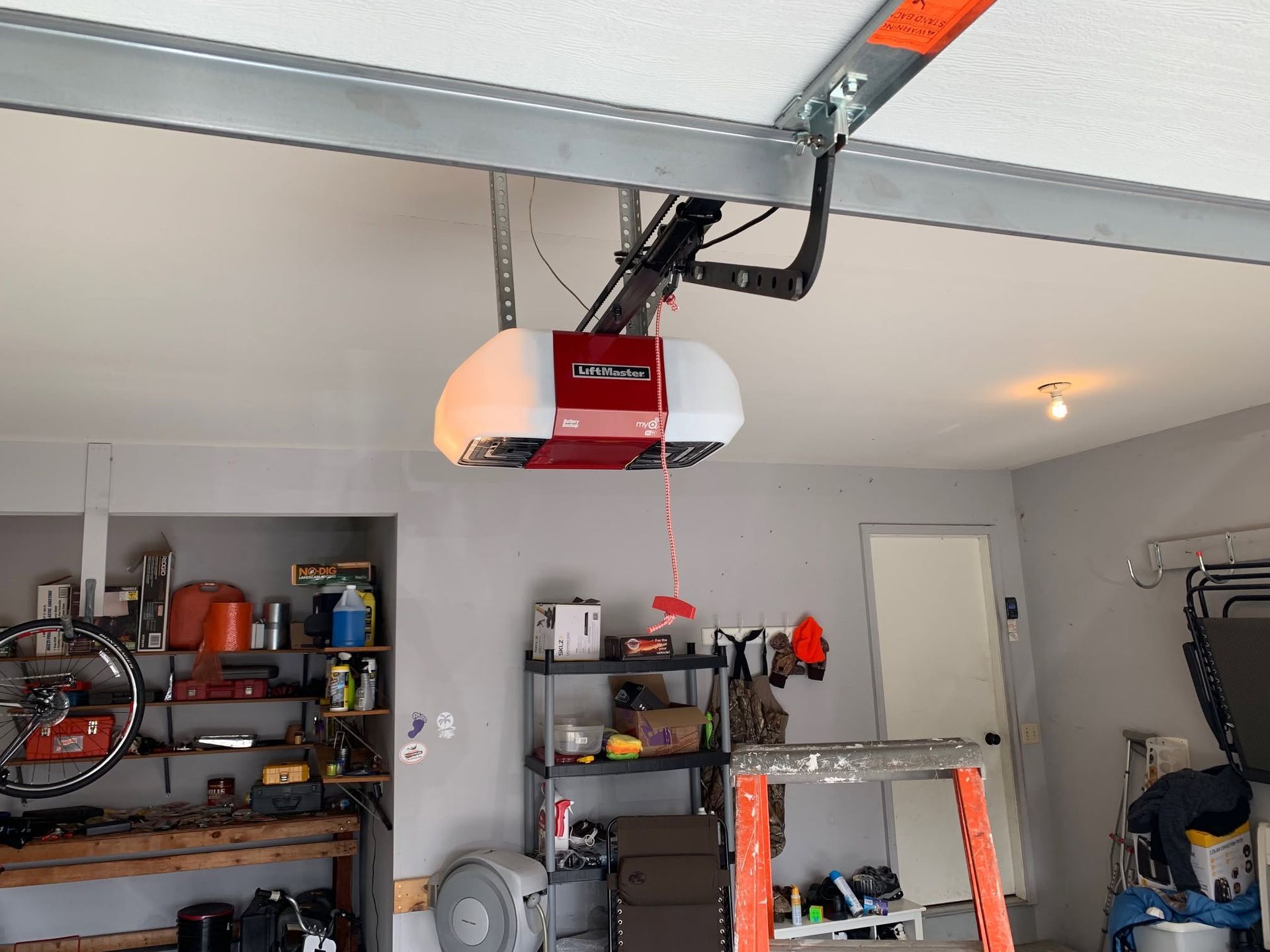 Red garage door opener mounted on a ceiling in a cluttered garage with shelves and tools.