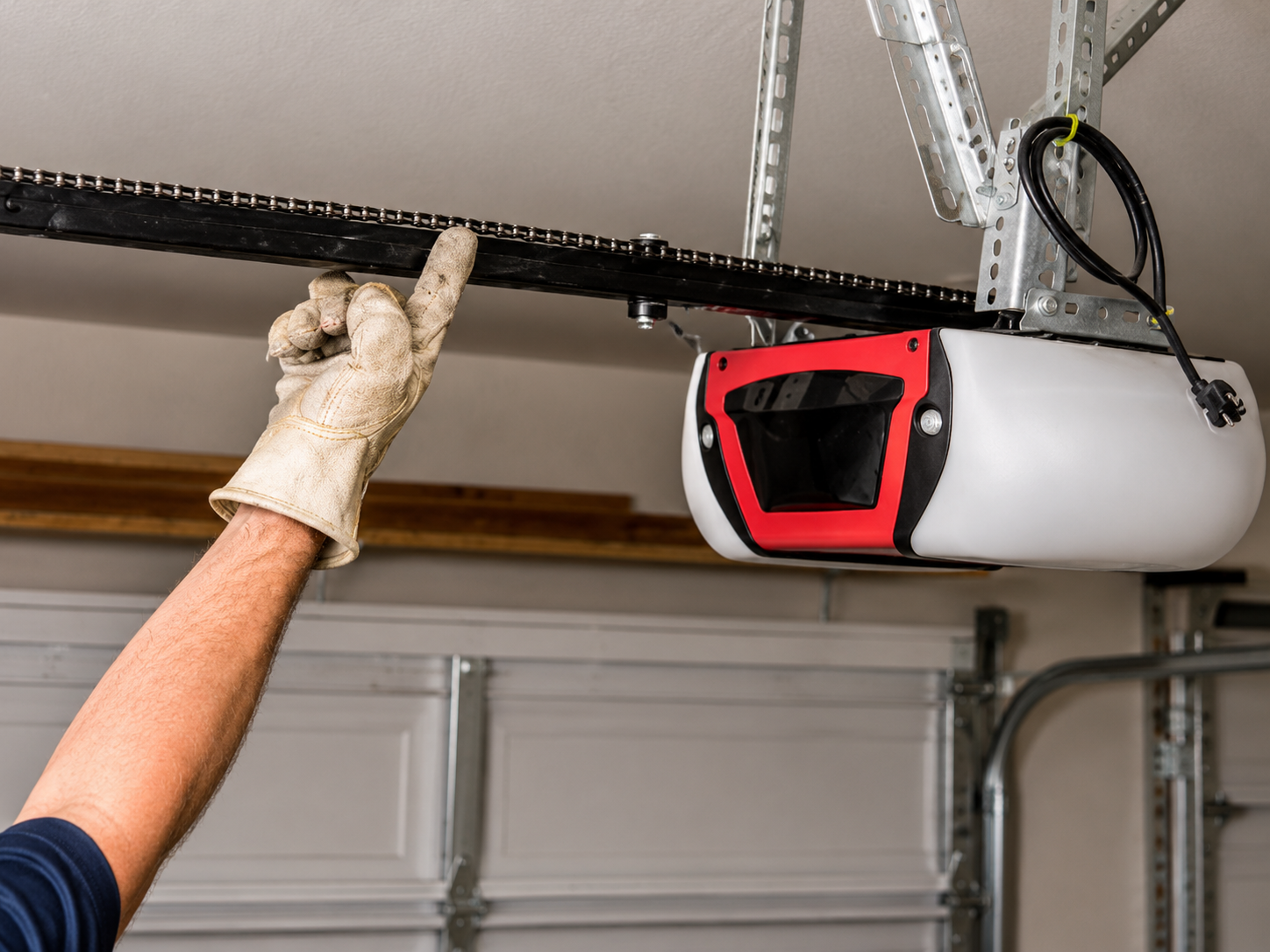 Gloved hand lifting a garage door opener rail near the ceiling above a white garage door