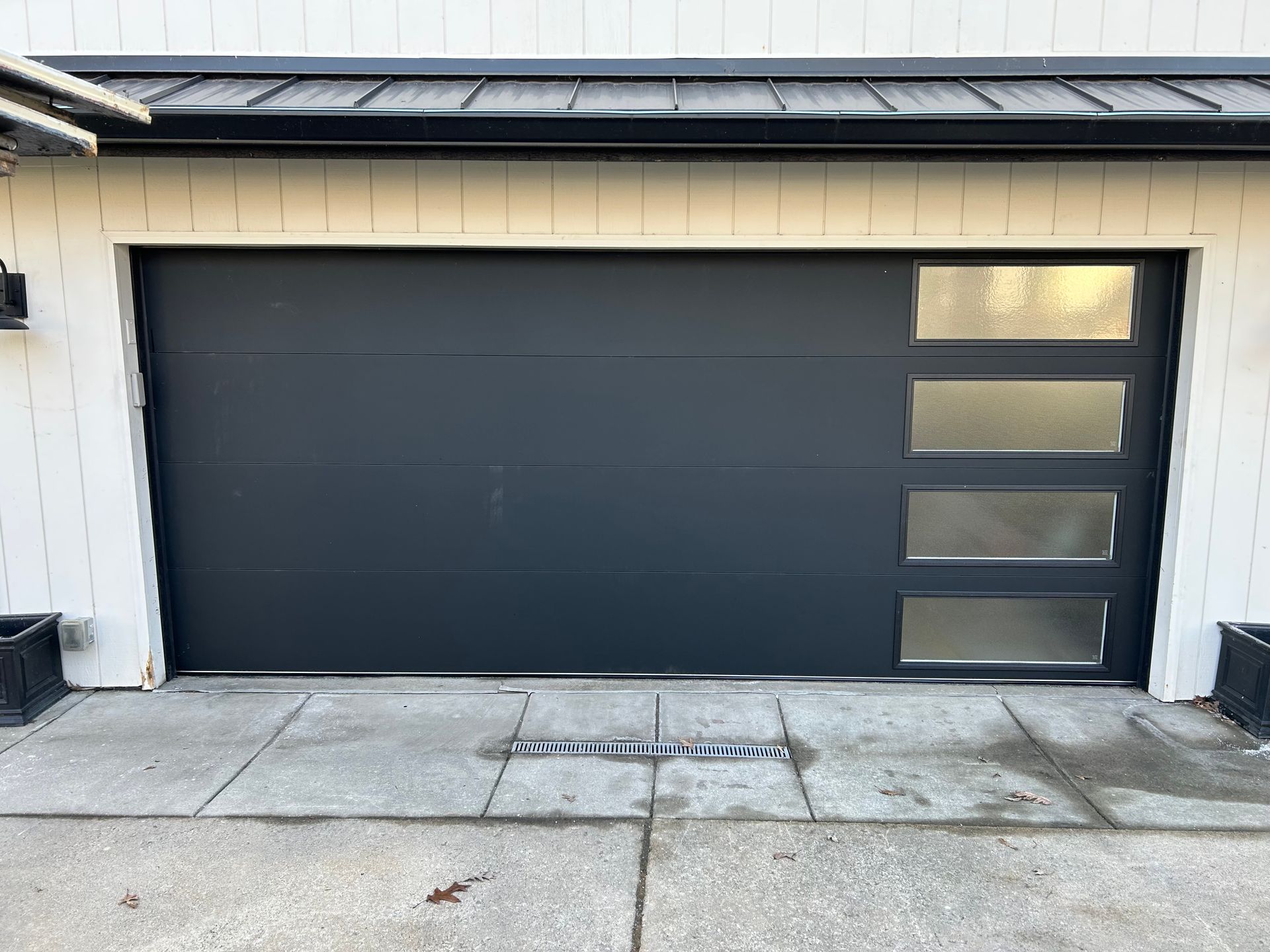 Modern black garage door with four frosted windows on the right, set in a white exterior wall.