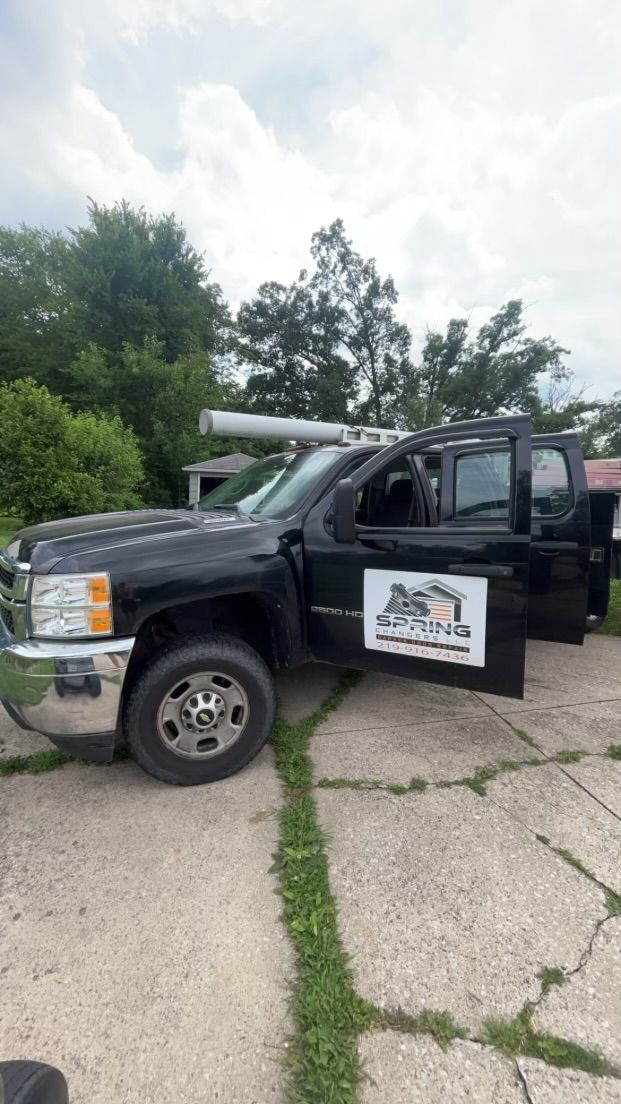 Black pickup truck with open door and logo on the side, parked on a cracked driveway near trees.