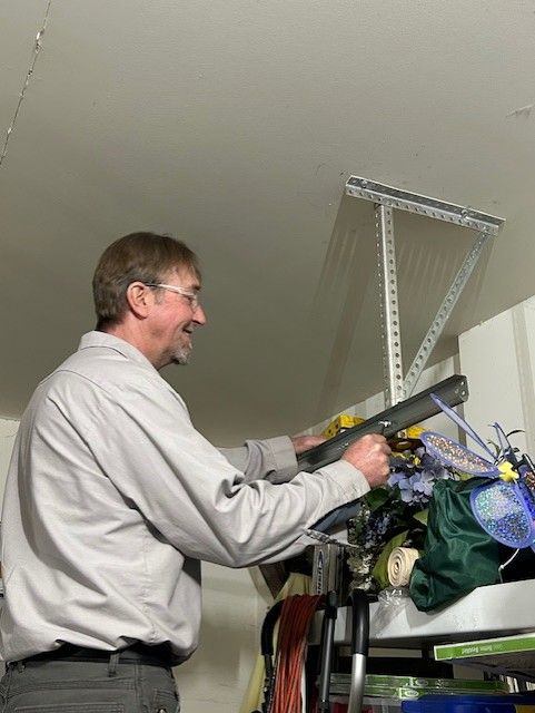 Man adjusting items on a high shelf in a storage room, reaching up beside boxes and flowers.