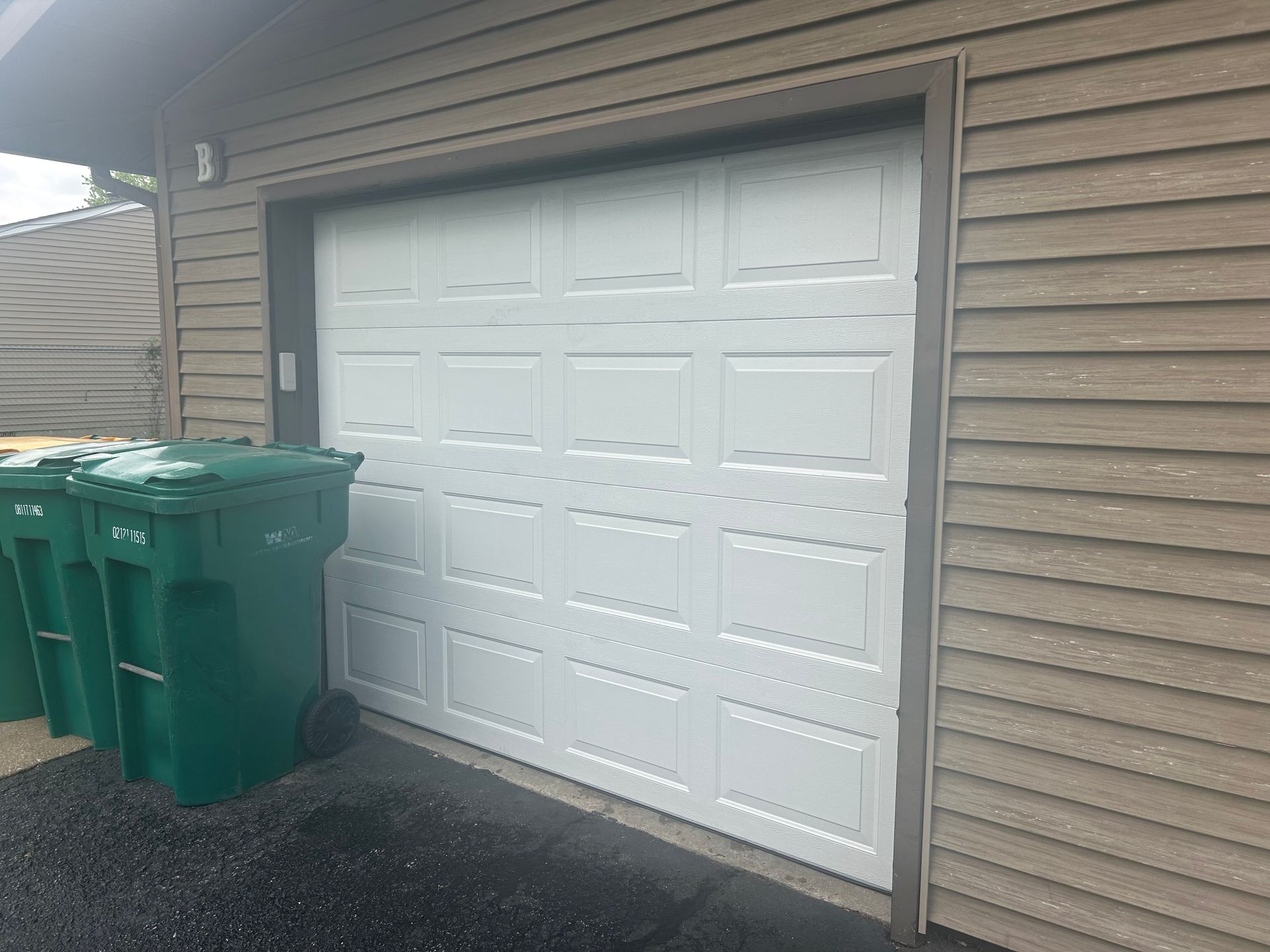 White garage door beside green trash bins against tan siding and wet pavement