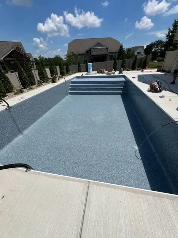 Empty swimming pool with blue speckled lining, concrete steps, and surroundings under a bright blue sky.