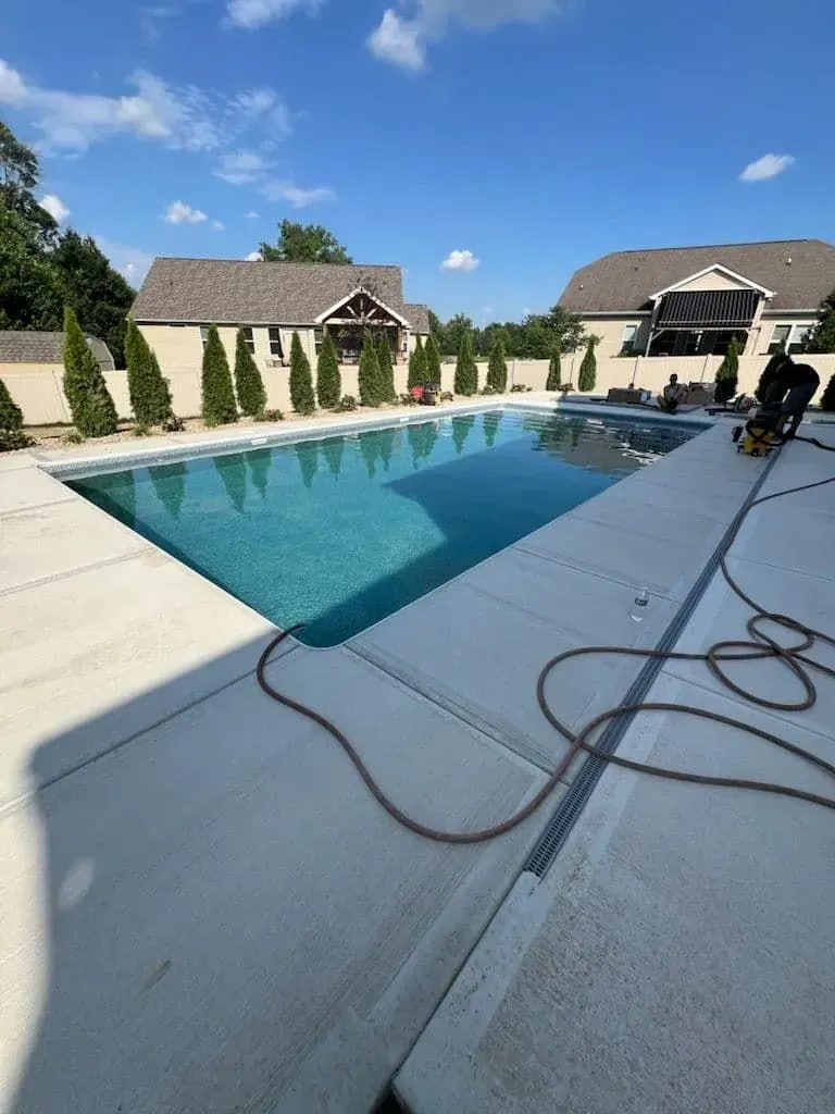 Swimming pool with turquoise water, surrounded by concrete and landscaping. Two houses in background under blue sky.