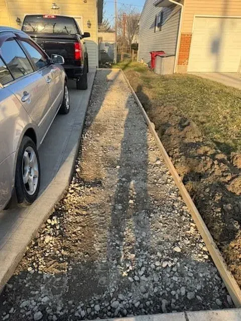 A narrow, gravel path with concrete borders in a residential driveway, next to grass and parked cars.
