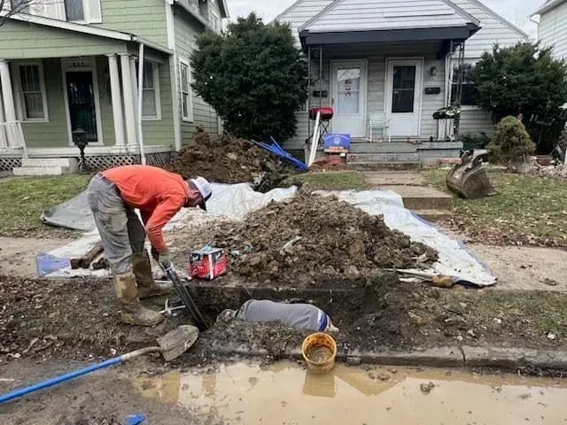 Person in a trench, inspecting a dark hole. Other person, gloves on, reaches into the hole. Mud and grass surrounding.