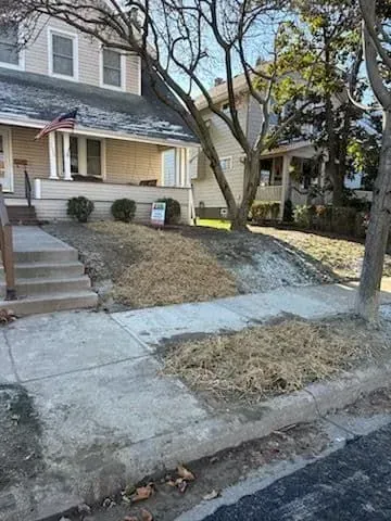 A two-story house with a porch. A sloped lawn is covered in straw and borders a sidewalk.
