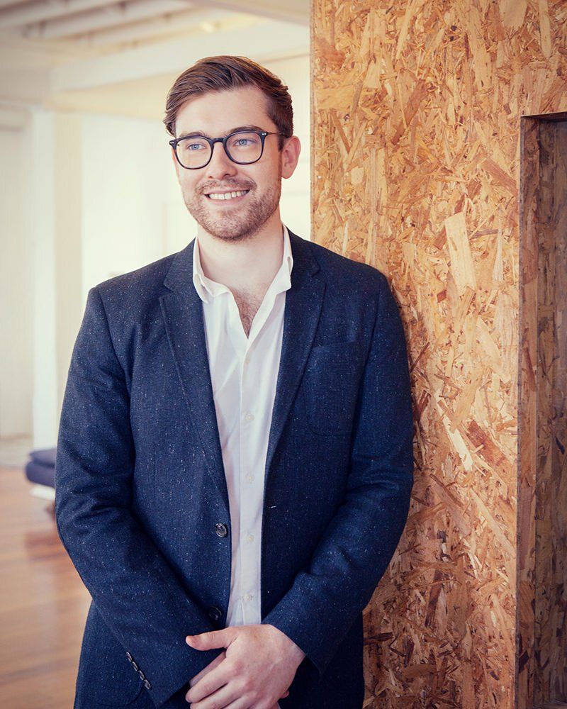 A man in a suit and glasses is leaning against a wooden wall.