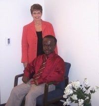 Woman in a red blazer stands behind a Black man seated in a chair, both smiling indoors. White flowers in the corner.