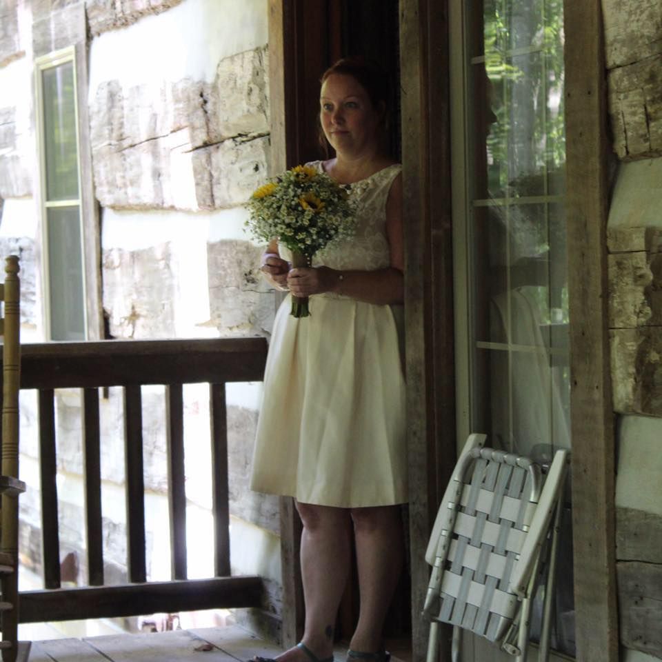 Woman in a white dress holding flowers stands on a porch. Rustic wood frame, folding chair nearby.